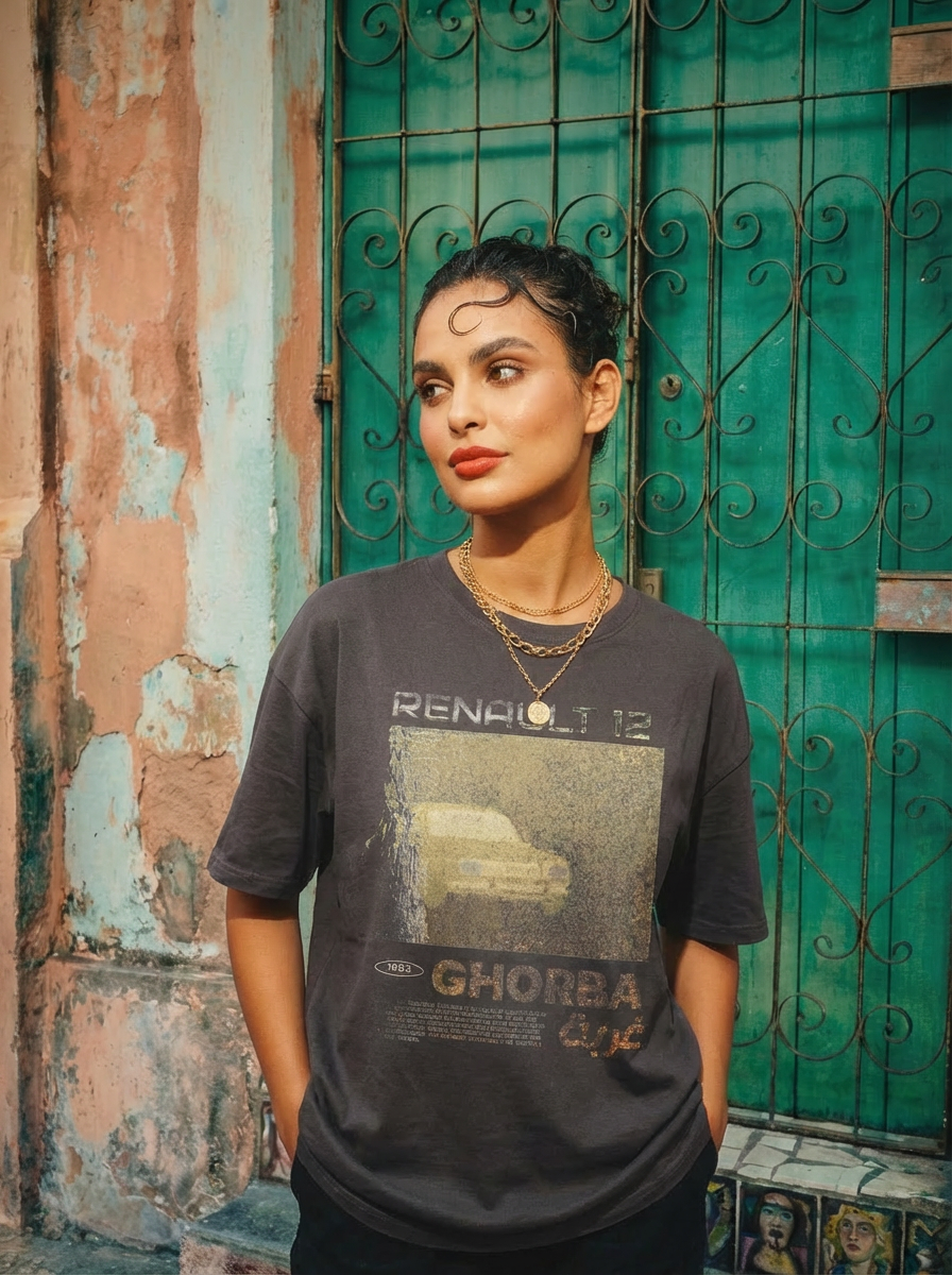 Une femme brune aux grands yeux se tient devant une porte verte, portant un t-shirt marron avec un motif Renault 12 effacé et l’inscription  GHORBA